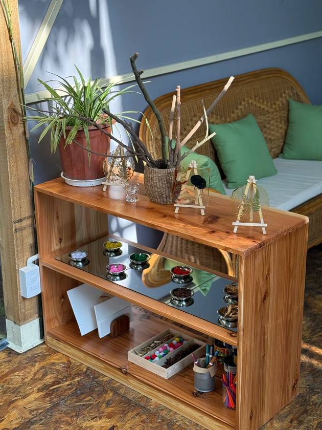 Wooden art shelf with pressed-flower frames and bowls of pompoms and spices, beside a rattan sofa