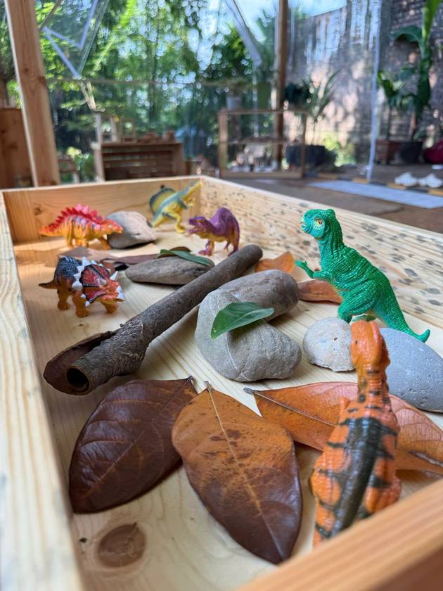 Toy dinosaurs among river stones, bark and fallen leaves in a wooden tray