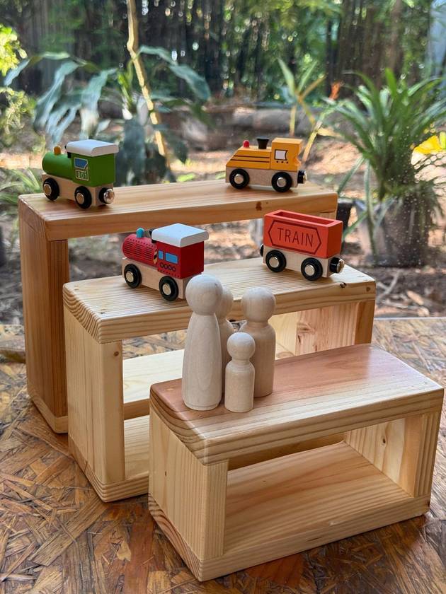 Colourful wooden trains and peg people on stepped wooden risers in front of the garden